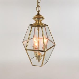May include: A vintage brass and glass pendant light fixture. The geometric-shaped lantern has clear glass panels and a brass frame. The fixture has three small light bulbs, and is suspended from a chain.