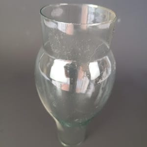 May include: A clear glass vessel featuring a wide, rounded upper body that tapers into a narrower, cylindrical base. The glass is transparent with a smooth surface, showing a distinct curved silhouette and a wide, open circular rim at the top.