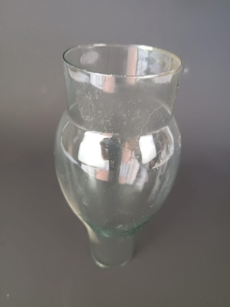 May include: A clear glass vessel featuring a wide, rounded upper body that tapers into a narrower, cylindrical base. The glass is transparent with a smooth surface, showing a distinct curved silhouette and a wide, open circular rim at the top.