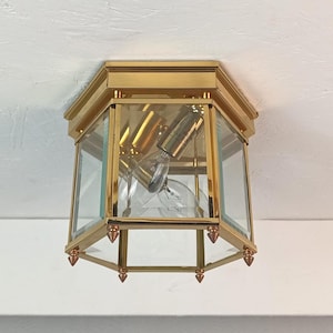 May include: A vintage ceiling light fitting with a gold-coloured metal frame and clear glass panels. The fitting has an octagonal shape and features a visible light bulb. The design is reminiscent of classic interior lighting.