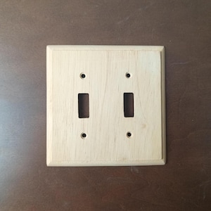 Puede incluir: Una placa de interruptor doble de madera de color claro con dos aberturas rectangulares para los interruptores y cuatro pequeños orificios circulares para el montaje. La placa tiene forma cuadrada con bordes ligeramente redondeados.