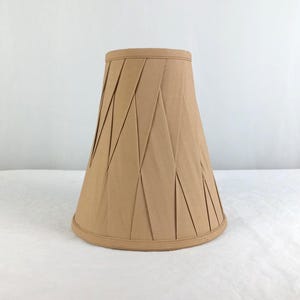 May include: A tan lampshade with a pleated design. The lampshade has a conical shape, with a wider base and a narrower top. The pleats create a geometric pattern, adding texture and visual interest. The shade is made of a fabric material.