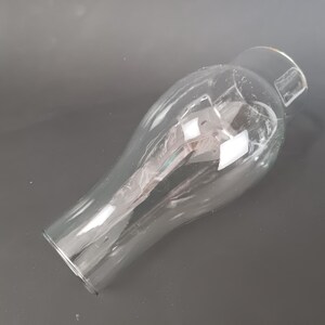 May include: A clear glass lamp shade with a bulbous, rounded body that tapers toward a narrower, cylindrical opening at the top. The glass is transparent and smooth, featuring a wide, open base and a reflective surface that catches light.