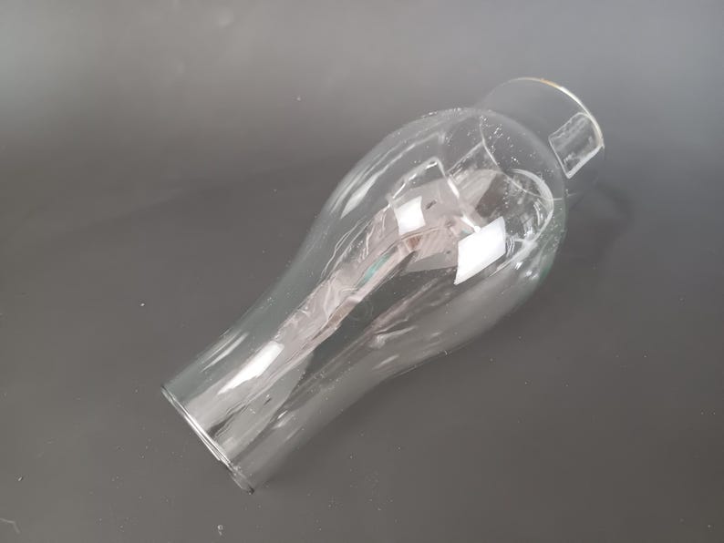 May include: A clear glass lamp shade with a bulbous, rounded body that tapers toward a narrower, cylindrical opening at the top. The glass is transparent and smooth, featuring a wide, open base and a reflective surface that catches light.