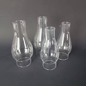 Puede incluir: Cuatro pantallas de lámpara de vidrio transparente de diferentes tamaños. Cada pantalla tiene un cuerpo redondeado y una parte superior acampanada, con una base circular. El vidrio transparente permite el paso de la luz. Están dispuestas sobre una superficie gris oscuro.