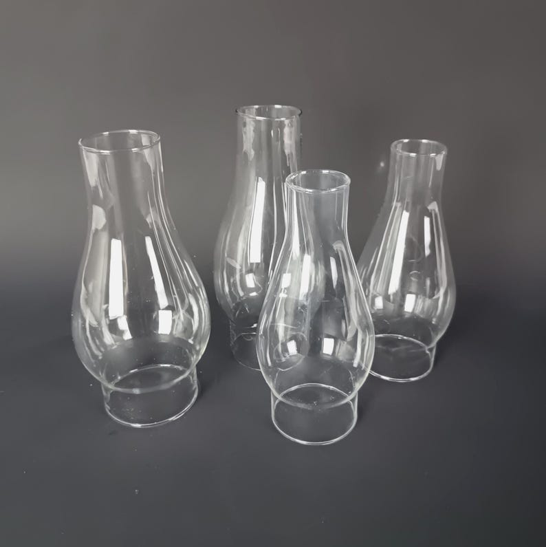 May include: A set of four clear glass lamp chimneys, each featuring a bulbous base that tapers into a narrow, cylindrical top opening. These transparent glass shades are designed for oil lamps and provide a smooth, reflective surface throughout.