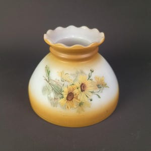 Peut inclure: Un abat-jour en verre en forme de cloche avec une base blanche et un dégradé de couleur jaune ambré en haut et en bas. La surface extérieure est décorée d'un motif floral peint représentant des tournesols jaunes avec des tiges et des feuilles vertes.