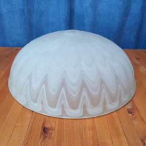 May include: A white, dome-shaped glass lampshade with a wavy, frosted pattern. The lampshade is sitting on a wooden surface. The lampshade is a light fixture component.