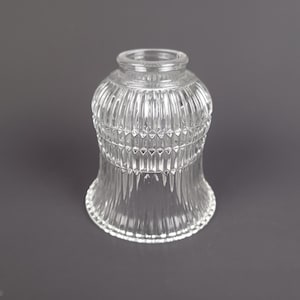 May include: A clear glass lampshade with a ribbed design. The lampshade has a flared shape and a rounded top. The glass is transparent, allowing light to pass through. The lampshade is designed to fit over a light bulb.