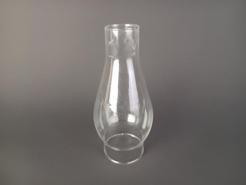 May include: A clear glass lamp chimney with a bulbous midsection that tapers toward a narrow top opening and a wider circular base. The transparent glass features a smooth, reflective surface and a simple, functional silhouette for light fixtures.
