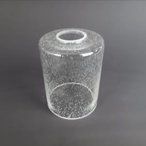 May include: A clear glass lampshade, cylindrical in form, with a small aperture at the top. The glass is textured with tiny, irregular bubbles. The lampshade is designed to diffuse light, suitable for a lamp.