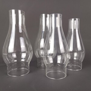 May include: A set of four clear glass lamp chimneys, each featuring a bulbous midsection that tapers toward a narrow top opening and a wider base. The transparent glass shades are arranged in a cluster, showcasing their smooth, curved silhouettes.