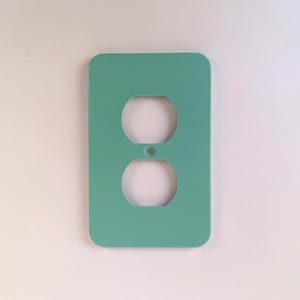 Op de afbeelding: Een turquoise rechthoekige stopcontactafdekking met afgeronde hoeken. Het heeft twee ronde openingen voor stekkers en een kleinere centrale opening. De achtergrond is wit. Ongeveer 12 cm hoog.
