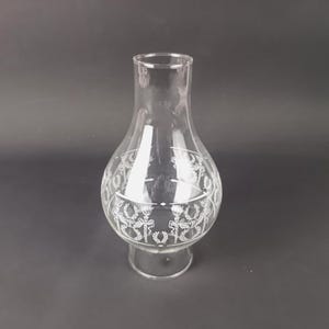 May include: Clear glass lamp shade with a decorative white floral pattern around the bulbous center. The shade has a flared base and a narrow top. The background is a solid dark gray.