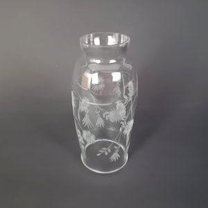 May include: A clear glass vase featuring an etched floral design of daisies and leafy stems. The vase has a rounded body, a slightly flared rim, and a smooth, transparent surface that showcases the delicate white engraved botanical pattern.