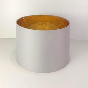 May include: A cylindrical drum lampshade featuring a smooth, light gray exterior fabric finish and a reflective, metallic gold interior lining. The shade includes a wire frame structure visible at the top opening for mounting to a lamp base.