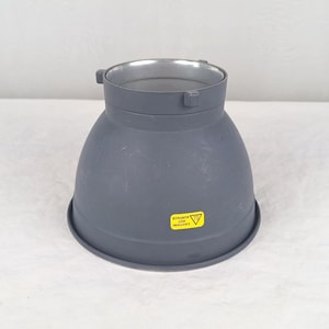 May include: A gray, cone-shaped metal object with a silver interior. The object has a wide base and a narrower top with two small tabs. A yellow sticker with the text "CAUTION! HOT SURFACE" is affixed to the side.