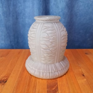 May include: A frosted glass lamp shade with a bulbous body and a flared base. The shade has a textured, wave-like pattern and vertical bands. The base has a ribbed design. The shade is sitting on a wooden surface.