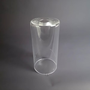 May include: A clear, cylindrical glass lamp shade featuring a smooth, transparent surface. The object has an open bottom and a smaller circular opening at the top, designed for light fixture installation. The glass is entirely colorless and reflective.