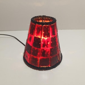 May include: A conical lamp shade constructed from interconnected translucent red square panels featuring a subtle star pattern. The top and bottom edges are finished with a border of small black beads, and a black power cord extends from the side.