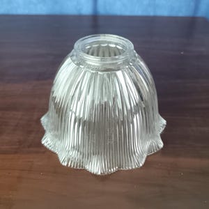May include: Clear glass lampshade with a fluted, bell-shaped design. The shade has a scalloped edge and a textured surface. The glass is transparent, allowing light to pass through. This lampshade is designed for use with a lamp.