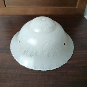 May include: A vintage, off-white glass lampshade with a scalloped edge. The shade features an embossed floral and leaf design, with a textured pattern on the top. The shade has a small hole for mounting and is likely from an antique lamp.