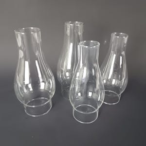 May include: A set of four clear glass lamp chimneys, each featuring a bulbous midsection that tapers toward a narrower top opening and a straight base. These transparent, hollow glass shades are designed for oil lamps and provide a clear view.