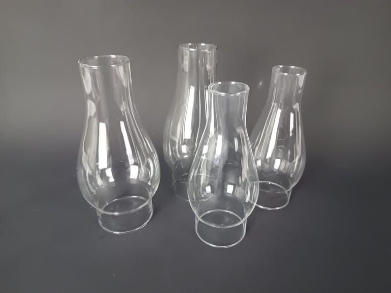 May include: A set of four clear glass lamp chimneys, each featuring a bulbous midsection that tapers toward a narrower top opening and a straight base. These transparent, hollow glass shades are designed for oil lamps and provide a clear view.
