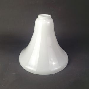 May include: A white glass lampshade with a rounded base and a flared, bell-shaped design. The lampshade has a smooth, glossy surface and a small opening at the top. The overall shape is reminiscent of a classic vintage design.