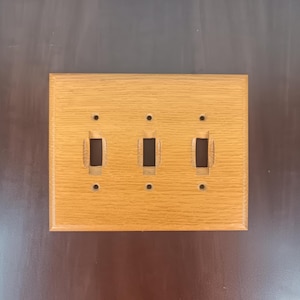 Puede incluir: Una placa de interruptor triple de madera marrón claro. La placa tiene tres aberturas rectangulares para los interruptores y tres pequeños agujeros para los tornillos de montaje. La veta de la madera es visible y la placa tiene un acabado liso.