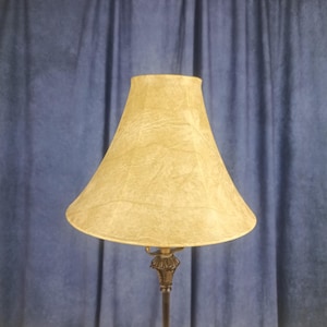 May include: A vintage-style lamp with a textured, light yellow lampshade. The lampshade is cone-shaped and sits atop a decorative, dark metal base. The background is a blue curtain.