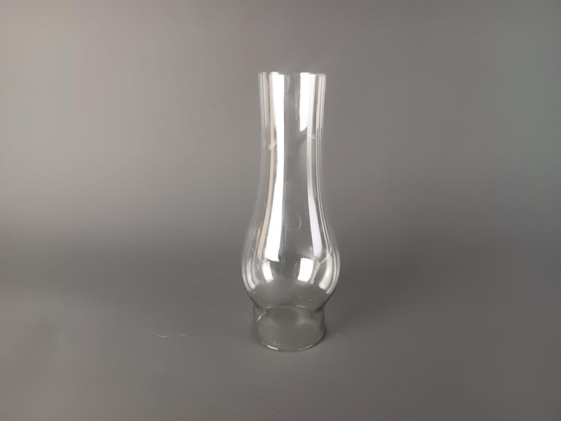 May include: A clear glass lamp chimney with a bulbous base that tapers into a slender, straight neck. The transparent glass features a smooth, reflective surface with a wide opening at the bottom and a slightly narrower opening at the top.