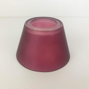 May include: A deep magenta-colored, frosted glass candle holder. The candle holder has a conical shape with a wide base and a narrow opening at the top. The glass has a smooth, matte finish.