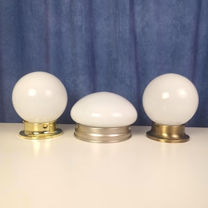 May include: Three vintage light fittings with white glass globes. Two are spherical, and one is a flattened dome shape. Each sits on a brass or gold-toned metal base. The background is a blue fabric.