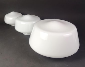 May include: Three white glass lamp shades arranged in a row, each featuring a smooth, rounded, dome-like shape with a small circular opening at the base. The shades vary in size, with the largest positioned in the foreground and two smaller ones behind.