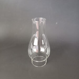 May include: A clear glass lamp chimney with a bulbous midsection and a straight, narrow neck. The glass is transparent and smooth, featuring a wider base that tapers inward before expanding into a rounded body, ending in a straight top opening.