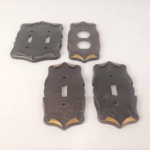 Può includere: Un set di quattro placche a muro marrone scuro con una finitura testurizzata e marmorizzata, arricchite da dettagli color oro sui bordi. La collezione include una placca per interruttore doppio, una per presa elettrica e due per interruttore singolo.