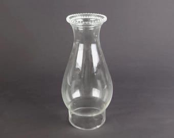 May include: A clear glass lamp chimney with a bulbous body and a flared top opening. The top rim features a decorative beaded texture, while the base is cylindrical and smooth, designed to fit securely onto a lamp fixture for light diffusion.