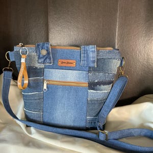 May include: A denim patchwork crossbody bag with a brown leather zipper pull and strap. The bag features a front pocket with a gold zipper and a brown leather tag that reads "Dona Dianna". The bag has a denim strap and a denim handle.