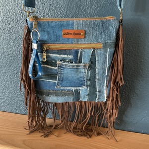 May include: A patchwork denim crossbody bag featuring various shades of blue fabric, brown leather fringe trim on the sides and bottom, a brown leather label with the text Dena Dianna, a gold zipper, and a blue keychain strap with white text.