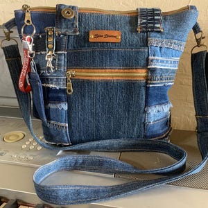 May include: A patchwork denim crossbody bag featuring various shades of blue fabric, a brown leather label with the text Dena Dianna, a front zippered pocket with brass hardware, a snap closure, and a red keychain tag with white text attached.