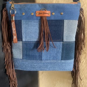 May include: Blue denim crossbody bag with a patchwork design. The bag features brown fringe on the sides and a brown leather tag with the text "Dena Dianna". A brown leather keychain with text hangs from the zipper.