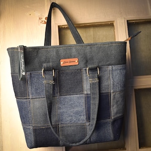 Puede incluir: Un bolso tote confeccionado con un patchwork de cuadrados de tela vaquera azul de diferentes tonos. El bolso cuenta con dos correas de hombro sujetas con herrajes color latón, una etiqueta de marca de cuero marrón con el texto "Dena Dianne" y un tirador de cremallera gris.