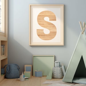 Peut inclure: Une affiche encadrée avec une grande lettre "S" à texture bois sur fond crème. L'œuvre est accrochée à un mur bleu clair au-dessus d'un sol en bois. Un petit tipi et diverses boîtes décoratives sont au premier plan.