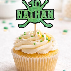 Peut inclure: Un cupcake avec un glaçage blanc et des vermicelles verts, surmonté d'un décor de gâteau vert à paillettes. Le décor de gâteau porte le chiffre 10 et le nom "NATHAN" dans un design sur le thème du hockey.