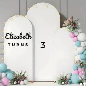 Peut inclure: Fond blanc en forme d'arche avec le texte "Elizabeth TURNS 3". La toile de fond est décorée de guirlandes lumineuses, de ballons bleus, roses et blancs, et d'arrangements floraux. Un design festif et célébratif.