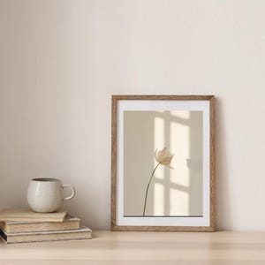 Peut inclure: Une impression d'art encadrée représentant une fleur beige délicate avec une longue tige sur un fond neutre. Le cadre en bois entoure l'œuvre, exposée sur une surface claire. Une pile de livres et une tasse sont à côté du cadre.