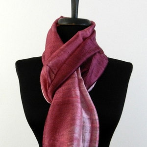 May include: A burgundy-colored silk scarf draped over a black mannequin. The scarf has a textured appearance with a gradient of colors from deep burgundy to a lighter pink. The scarf is lightweight and has fringed edges.