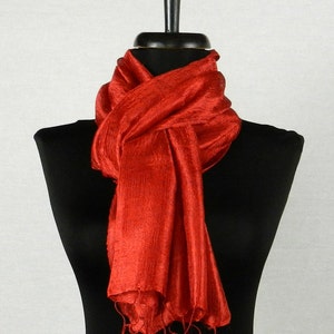 May include: A vibrant red scarf displayed on a black mannequin. The scarf is made of a silky material, with a textured appearance and fringed edges. The scarf is draped around the mannequin's neck, showcasing its flowing design.