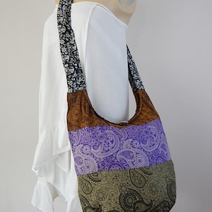 Puede incluir: Bolso de hombro patchwork con estampado de cachemira en marrón, morado y verde oliva. La correa es negra con un diseño floral blanco. El bolso se exhibe en un maniquí con un vestido blanco.